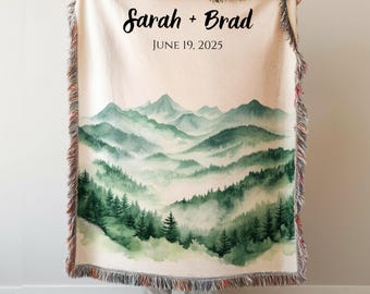 May include: A cream-colored throw blanket featuring a watercolor painting of a mountain landscape in shades of green and gray. The blanket has fringed edges and the text "Sarah + Brad" and "JUNE 19, 2025" in black script.