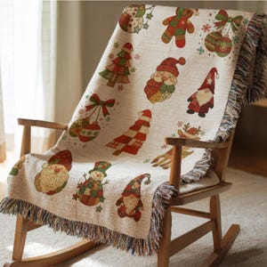 May include: A cream-colored Christmas throw blanket draped over a wooden rocking chair. The blanket features festive illustrations of Santa Claus, snowmen, gnomes, Christmas trees, ornaments, and snowflakes. The blanket has a fringed edge.