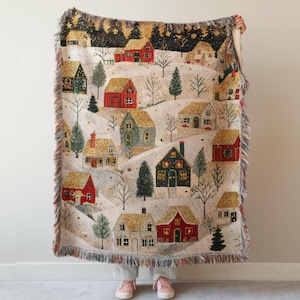 May include: A woven throw blanket featuring a winter village scene with red, green, and gold houses nestled in a snowy landscape. The design includes evergreen trees, bare branches, and a dark night sky with stars, finished with a fringed edge.