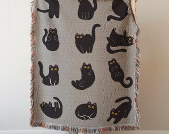 May include: A gray throw blanket featuring a repeating pattern of black cat illustrations with yellow eyes and purple accents. The blanket has a fringed edge with a mix of colors. The cats are in various poses.