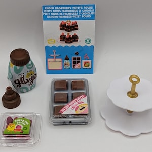 Peut inclure: Plusieurs miniatures alimentaires, dont une bouteille de "glaze", un plateau de carrés de chocolat et un présentoir blanc à deux niveaux. Un paquet de friandises "Happy Sun Farms" et une carte avec le texte "Choco Raspberry Petits Fours" sont également présents.
