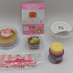 Peut inclure: Un ensemble de fournitures miniatures pour la décoration de gâteaux, comprenant un gâteau rose et blanc, un support de gâteau blanc, une assiette à gâteau blanche avec un couteau argenté, un petit récipient de glaçage rose et un paquet de vermicelles roses et blancs. Le paquet de vermicelles porte l'inscription "Les Sun-Baked Sweets Ladybug Cake".