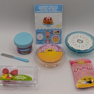 Peut inclure: Un ensemble de fournitures miniatures pour la fabrication de gâteaux, comprenant un récipient bleu étiqueté "Vanilla", un gâteau éponge jaune, un paquet rose étiqueté "Crumble", une assiette bleue avec un motif blanc et un récipient en plastique transparent avec des fraises rouges et blanches.