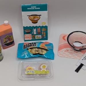 Peut inclure: Un ensemble de nourriture ramen miniature avec divers composants. Comprend une boîte avec une illustration de bol de ramen, un paquet de nouilles, un petit récipient d'assaisonnement, un bol, une cuillère, des baguettes et un petit récipient avec des œufs.