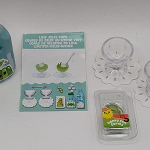 Peut inclure: Un ensemble de coupes de gelée au citron vert, de crème fouettée et d'une boisson aigre au citron vert. L'emballage présente un citron vert de dessin animé avec un visage souriant. Le texte sur l'emballage indique "Lime Jelly Cups", "Coupés de Gelée au Citron Vert", "Copas de Gelatina de Lima", "Limetten-Gelez-Becher", et "Happy Sun Farms".