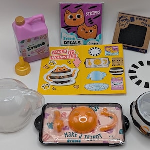 May include: A set of craft supplies for making a teapot. The set includes a clear plastic mold, a pink plastic bottle, a yellow plastic teapot, a sheet of stickers, and a sheet of instructions. The instructions say "Make a Teapot Studio".