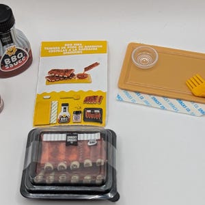 May include: A miniature set of BBQ grilling supplies, including a bottle of BBQ sauce, a jar of BBQ spice rub, a tray with a small bowl and a yellow fork, and a pack of ribs.
