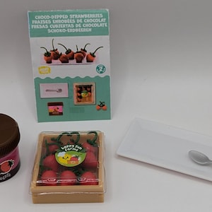 Peut inclure: Un ensemble de miniatures d'aliments pour une maison de poupée, comprenant une boîte de fraises enrobées de chocolat, un pot de pâte à tartiner au chocolat, une assiette blanche avec une cuillère en argent et une carte avec le texte "CHOCO-DIPPED STRAWBERRIES FRAISES ENROBÉES DE CHOCOLAT FRESAS CUBIERTAS DE CHOCOLATE SCHOKO-ERDBEEREN".