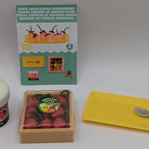 Peut inclure: Un ensemble miniature de nourriture en plastique, comprenant une boîte de fraises rouges, une assiette jaune avec une cuillère en argent et un petit pot de sauce au chocolat. La boîte porte l'inscription "Happy Sun Farms". L'emballage des fraises porte l'inscription "Fraises enrobées de chocolat blanc".