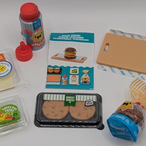 Peut inclure: Un ensemble de nourriture en plastique miniature, comprenant un steak de burger végétarien, du fromage cheddar blanc, une bouteille de ketchup et un pain. Les articles sont emballés dans des contenants en plastique transparent avec des étiquettes colorées. Les étiquettes présentent des personnages de dessins animés et du texte qui dit "Happy Sun Farms" et "Shacky Snacks Co."