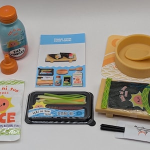 Peut inclure: Un kit de fabrication de sushis miniature avec divers composants. Comprend une bouteille de vinaigre de riz, un paquet de riz, un plateau de légumes, des algues nori, un bol avec une cuillère et une paire de baguettes. Le kit a une esthétique ludique.