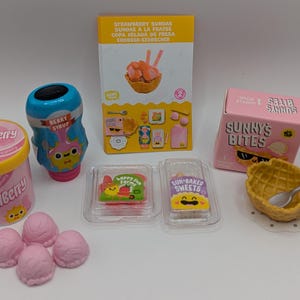 Peut inclure: Un ensemble de jouets miniatures sur le thème de la crème glacée, comprenant un contenant de crème glacée à la fraise, un cornet de gaufres, une bouteille de sirop de fraise et deux petits contenants de garnitures pour crème glacée. Les jouets sont tous roses et jaunes avec des visages de dessins animés et le texte "Strawberry", "Sunny's Bites", "Berry Strup", "Happy Fun Snacks", et "Sun-Baked Sweets".