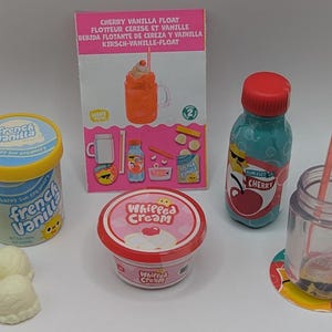 Peut inclure: Ensemble de jouets alimentaires, comprenant un pot de glace à la vanille française, une bouteille de soda à la cerise, un pot avec une paille et un pot de crème fouettée. Un emballage avec le texte "Cherry Vanilla Float" est également présent.