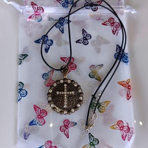 May include: A bronze-toned pendant necklace with a cross design, encrusted with clear rhinestones, hangs on a black cord. The necklace rests on a white butterfly-printed drawstring bag. The bag features butterflies in various colors.