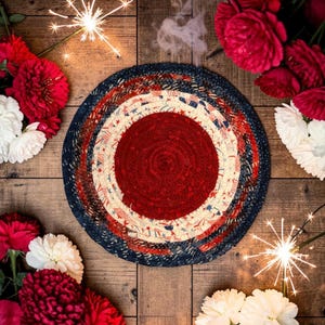 May include: A round, patriotic-themed rug with a red center, surrounded by concentric rings of red, white, and blue fabric. The design incorporates stars and stripes. The rug is displayed on a wooden surface, with flowers and sparklers.