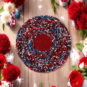 May include: A round braided rug with a patriotic color scheme. The rug features a central dark red circle surrounded by concentric rings of red, white, and blue braided fabric. The rug is on a wooden surface, surrounded by red flowers.