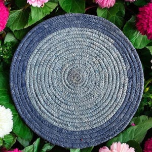 May include: A round, blue and gray woven placemat with a circular design. The placemat is surrounded by pink flowers.