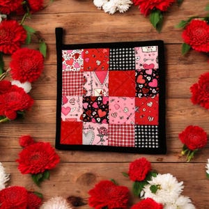 May include: A red and black patchwork pot holder with a heart and arrow design. The pot holder is surrounded by red and white flowers on a wooden background.