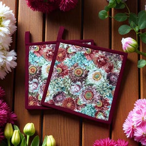 Könnte beinhalten: Zwei quadratische Topflappen mit einem Blumenmuster in Rot-, Burgunder- und Grüntönen auf hellblauem Hintergrund. Die Topflappen haben burgunderfarbene Ränder und Aufhängeschlaufen. Das Bild ist auf einer Holzoberfläche platziert, umgeben von Blumen.