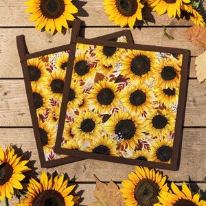 May include: Two square quilted pot holders featuring a vibrant sunflower pattern with yellow petals and dark brown centers, accented by small brown leaves and white floral details. Each pot holder has a solid dark brown fabric border and a hanging loop.