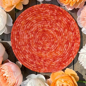May include: A circular, coiled rope mat featuring a vibrant, mottled pattern of orange, red, and yellow hues. The mat is constructed from tightly wound fabric cord, creating a textured, spiral surface design that is flat and round in shape.