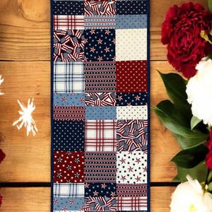 May include: A rectangular patchwork table runner with a navy blue border. The runner features squares with patriotic patterns, including American flags, stars, and plaid designs in red, white, and blue. The runner is displayed on a wooden surface.