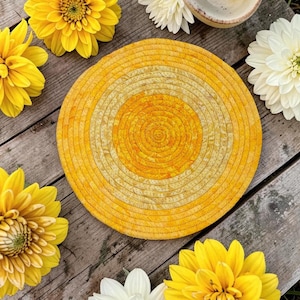Puede incluir: Un posavasos o salvamanteles circular de tela con un diseño de cuerda enrollada en un degradado de tonos amarillos y naranjas. La superficie textil muestra un patrón moteado y texturizado a lo largo de los círculos concéntricos.