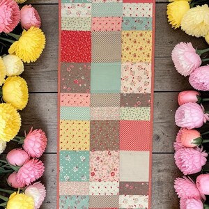 May include: A patchwork table runner with a variety of floral and geometric patterns in red, pink, yellow, and green. The runner is surrounded by pink and yellow flowers on a wooden surface.