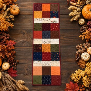 May include: A rectangular patchwork table runner featuring a grid of squares in various autumn-inspired colors and patterns, including orange, deep red, navy blue, olive green, and cream, finished with a solid dark red fabric border.
