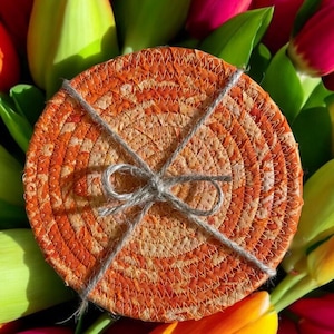 May include: A set of four orange and cream woven coasters tied together with twine. The coasters are round and have a textured surface. The coasters are surrounded by a bouquet of tulips.