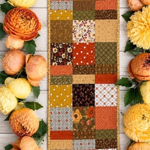Könnte beinhalten: Ein rechteckiger Tischläufer mit einem Patchwork-Design aus verschiedenen Stoffquadraten in Orange-, Gelb-, Braun- und Grüntönen. Die Muster umfassen Blumendrucke, kleine Punkte und Karotexturen, die alle von einer dünnen goldfarbenen Borte gesäumt sind.