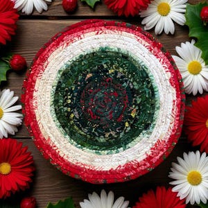 May include: A round, handcrafted trivet with a spiral design in red, white, and green. The trivet is surrounded by red and white daisies and holly on a wooden surface. The trivet's design is reminiscent of a holiday theme.