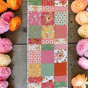 May include: A rectangular table runner featuring a patchwork design of various fabric squares. The fabric patterns include floral prints, geometric shapes, and solid colors in shades of pink, green, yellow, orange, and white, arranged in a grid.