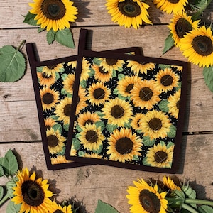 May include: Two square pot holders featuring a sunflower design on a black backdrop, edged with a dark brown border. The image is set on a wooden surface and surrounded by fresh sunflowers with yellow petals and dark brown centres.