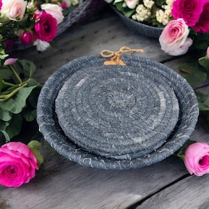 May include: Two gray woven rope baskets with a white rope trim. The baskets are stacked on top of each other. The baskets are on a wooden surface with pink flowers.