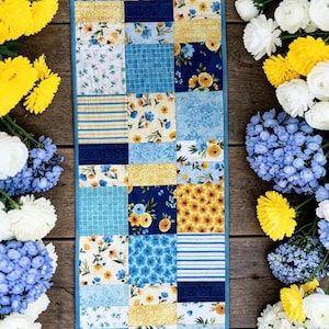 Pode incluir: Um caminho de mesa de patchwork com uma borda azul, apresentando quadrados de padrões florais e listrados em tons de azul, amarelo e branco. O caminho de mesa é cercado por buquês de flores amarelas, brancas e azuis em uma superfície de madeira.