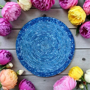 May include: A round, blue woven placemat with a spiral design is surrounded by vibrant peonies in shades of pink, yellow, and white. The flowers are arranged on a light gray wooden surface, creating a colorful and natural aesthetic.