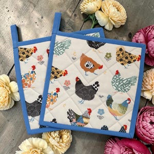 May include: Two square quilted oven mitts with a cream background featuring a pattern of colourful chickens and flowers. They have a blue border and a blue hanging loop. The oven mitts are on a wooden surface.