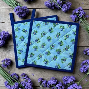 May include: Two square, quilted pot holders with a light blue floral pattern and dark blue trim. Each pot holder has a dark blue loop for hanging. The pot holders are surrounded by sprigs of purple flowers.