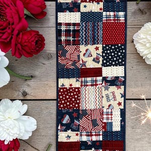 Könnte beinhalten: Ein rechteckiger, gesteppter Tischläufer mit einem Patchwork-Design aus roten, weißen und blauen Stoffquadraten. Die Muster umfassen Sterne, Streifen, Karos und Herzen, wobei ein Quadrat deutlich den Text Stars and Stripes in Rot und Blau zeigt.