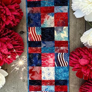 May include: A patriotic table runner with a patchwork design featuring red, white, and blue patterns, including stars and stripes. The runner is surrounded by red and white peonies, and a sparkler.