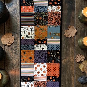 May include: A rectangular patchwork table runner featuring a variety of fabric squares with Halloween-themed patterns including pumpkins, black cats, moons, stars, and stripes in orange, black, purple, and white colors with a black border.