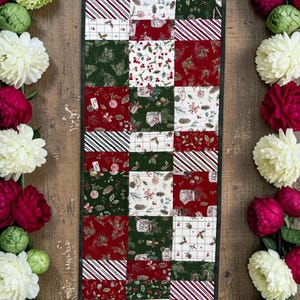 May include: A rectangular patchwork table runner featuring a grid of squares with red, green, and white holiday-themed patterns, including candy cane stripes, holly berries, and pine cones, flanked by two vertical garlands of white and red flowers.