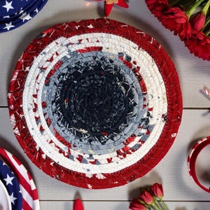 Könnte beinhalten: Ein runder, geflochtener Tischset in Rot, Weiß und Blau mit Sternenmuster. Das Tischset hat einen Durchmesser von etwa 30 cm. Das Design erinnert an patriotische Themen mit einem Spiralmuster in der Mitte.