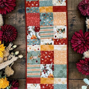 May include: A rectangular quilted table runner featuring a patchwork design of fabric squares in shades of sage green, mustard yellow, and deep red. The fabric patterns include floral prints, stripes, and solid colors, all bordered by a thin red trim.