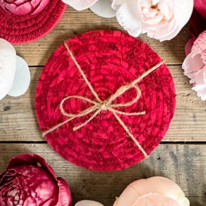May include: A set of red fabric coasters tied with twine, surrounded by peonies in shades of red, pink, and white. The coasters have a woven texture and are circular in shape. The background is a rustic wooden surface.