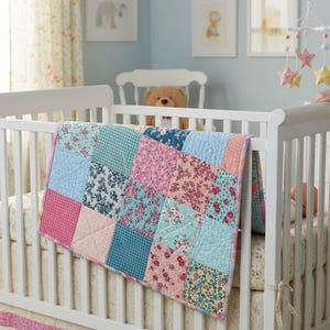 Könnte beinhalten: Ein weißes Babybett mit einer bunten Patchwork-Decke. Die Decke zeigt Quadrate aus rosa, blauen und türkisfarbenen Stoffen mit Blumenmustern. Über dem Bett hängt ein Mobile mit sternförmigen Ornamenten.