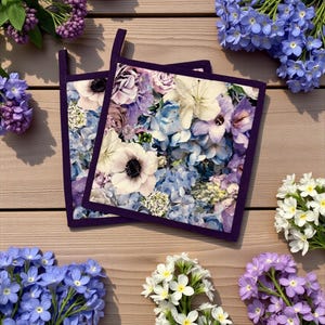 Könnte beinhalten: Zwei quadratische Topflappen mit einem Blumendesign mit weißen Anemonen, blauen Hortensien und lila Akzenten. Die Topflappen haben einen dunkelvioletten Rand und eine Schlaufe zum Aufhängen. Das Bild ist von frischen Blumen umgeben.