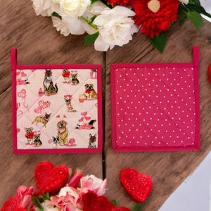 Könnte beinhalten: Zwei rosa und weiße gepolsterte Topflappen mit einem rosa Tupfenmuster auf der einen Seite und einem Muster von Hunden mit Herzen und Ballons auf der anderen Seite.