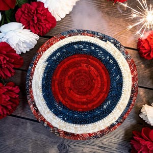 May include: A round, patriotic-themed coaster with concentric circles of red, white, and blue fabric. The coaster is surrounded by red and white flowers and a lit sparkler, suggesting a festive occasion.